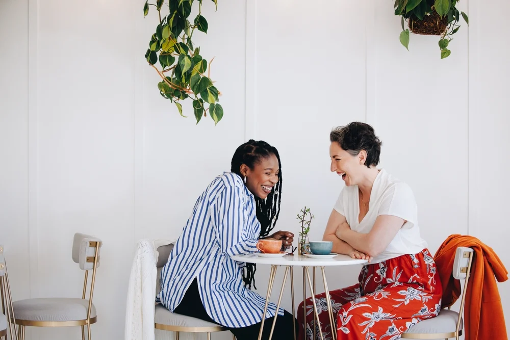 Two diverse female friends laughing together in a light, beautiful coffee shop