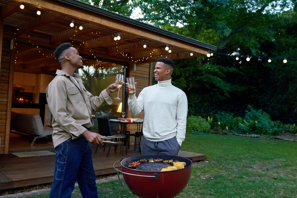Two happy African-american men by the barbeque making a toast in a gorgeous backyard with pendant lights