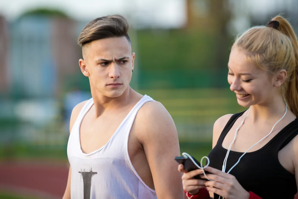 Jealous worried caucasian man peering over the shoulder of his girl friend while she is texting on the phone smiling