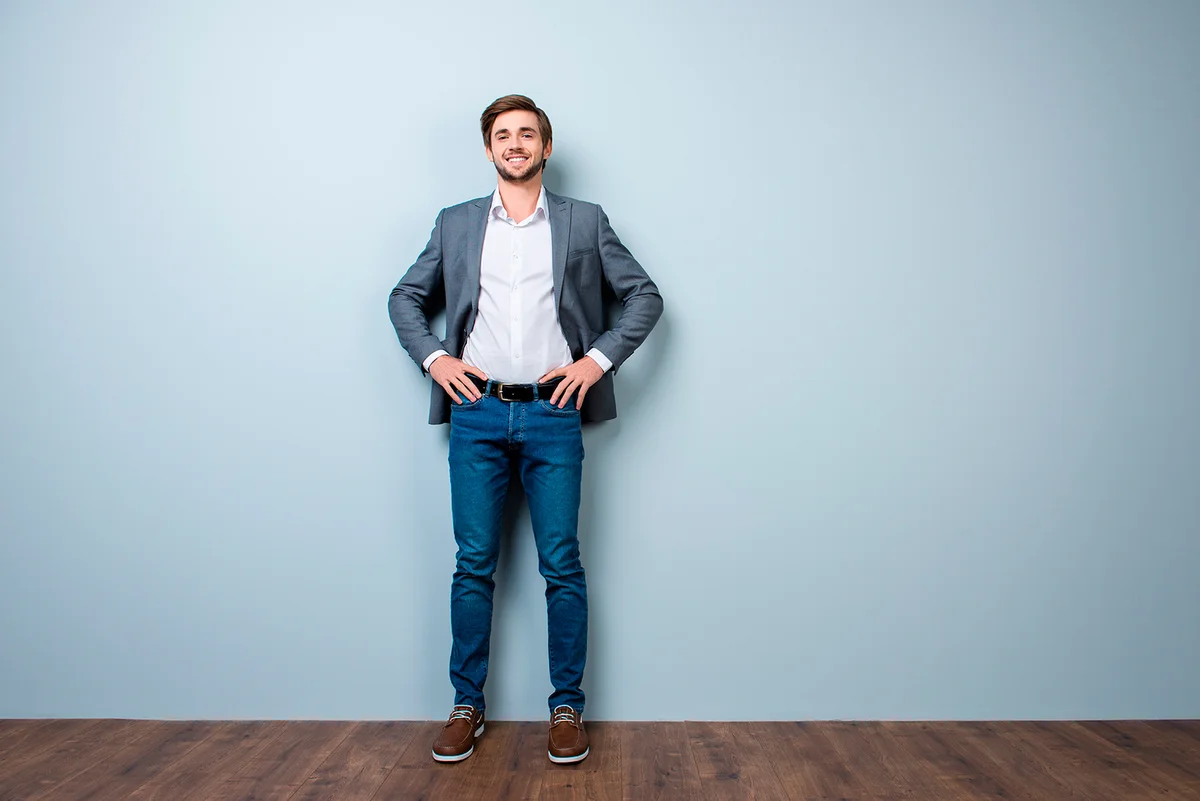Full-length photo of successful handsome young man in formal wear smiling while standing near gray wall.