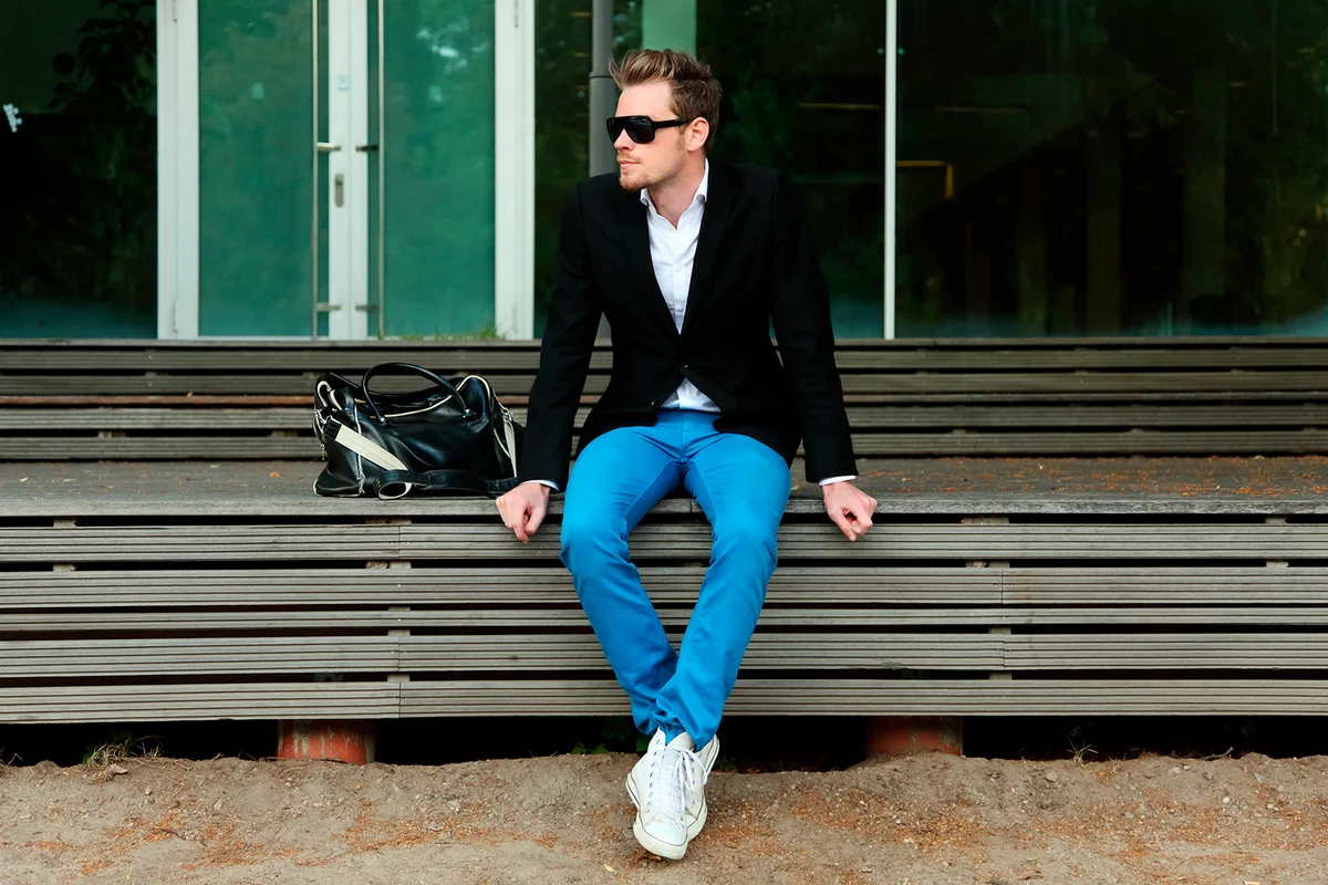 A man sitting outside wearing black sunglasses and a black jacket with blue jeans.