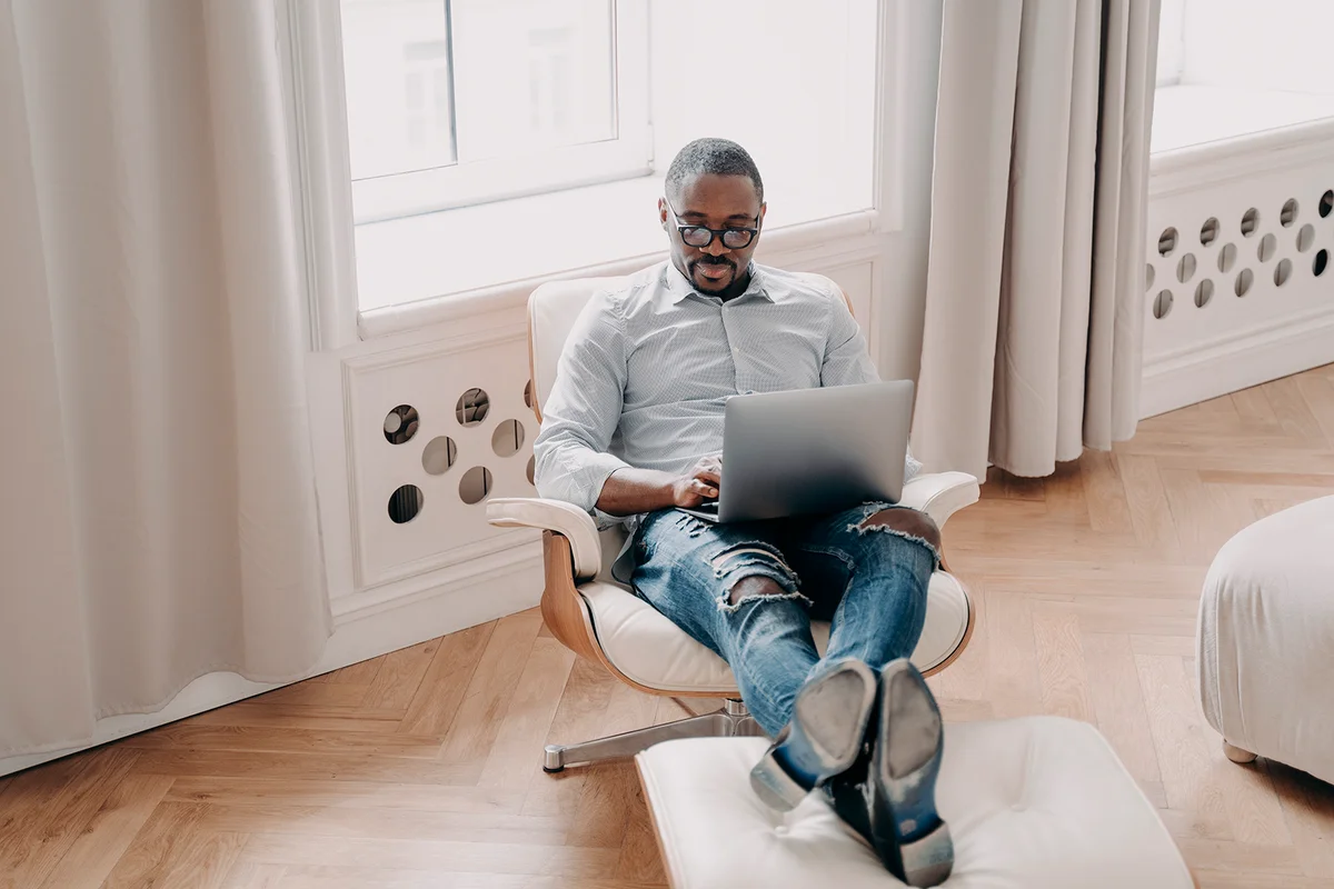 Focused african american businessman works online at laptop, sitting in armchair indoors. Modern black male employee manager in glasses answers email, developing business project. Remote job concept.