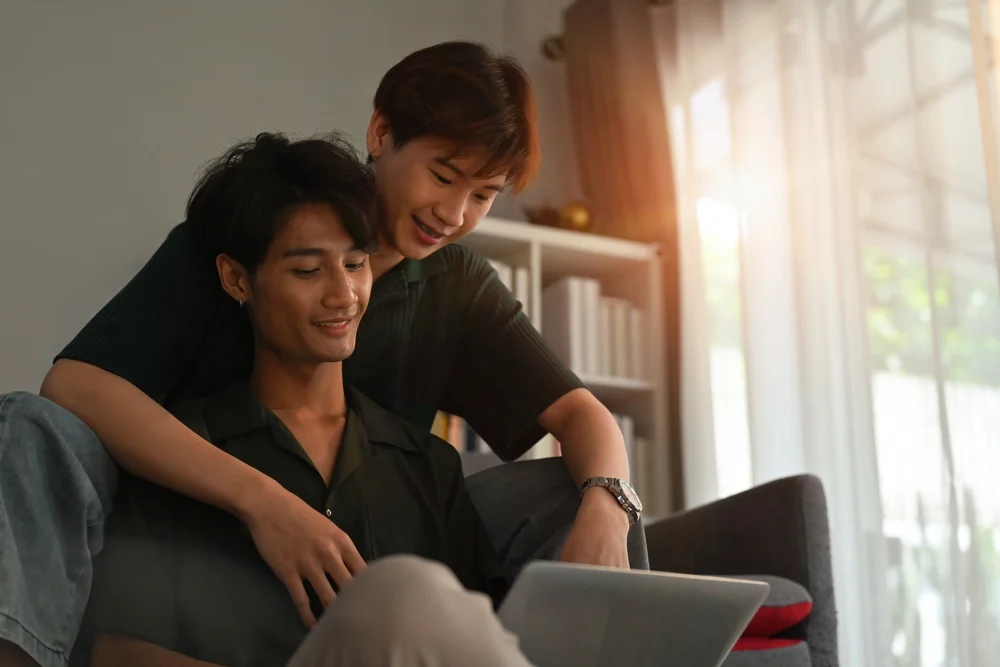 Young asian gay couple browsing laptop while sitting in the living room very close to each other