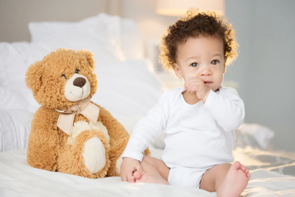 Cute baby boy with curly hair sitting in the bed by his teddy bear