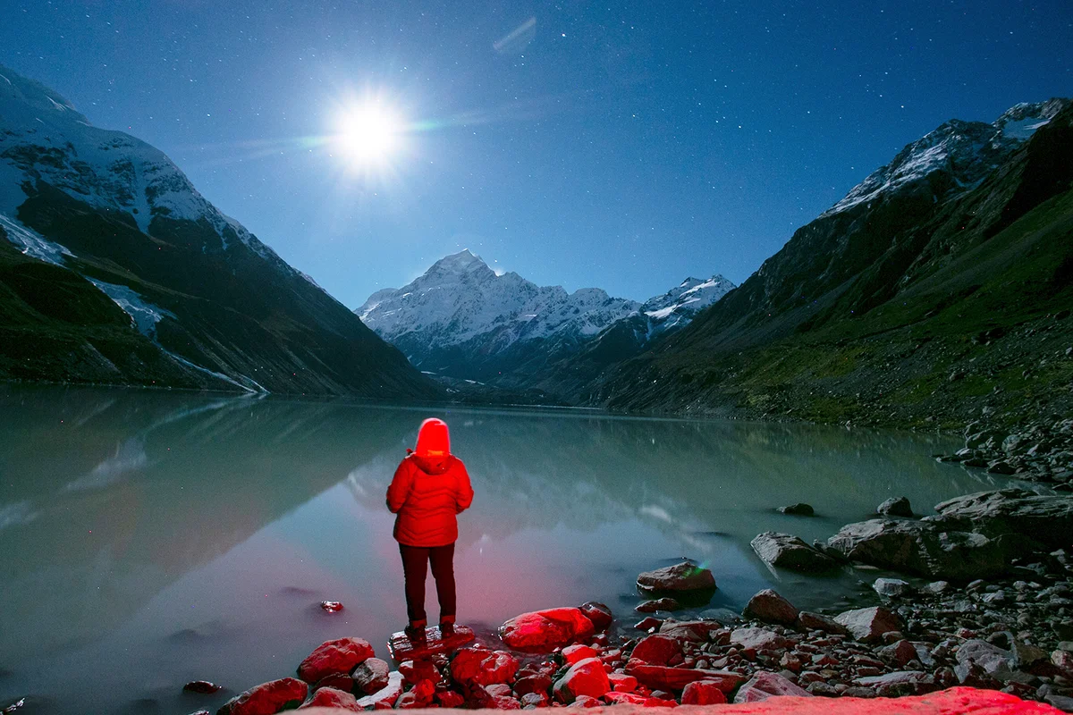 A person in a red jacket stands at the edge of a quiet mountain lake under a starry night sky, with snowy peaks reflected in the calm water. The full moon projects a serene glow over the landscape