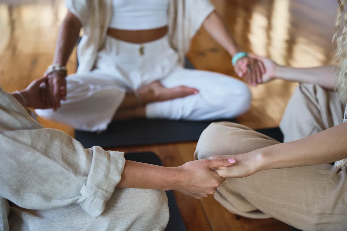 Group of conscious and healthy women sitting in a circle meditating together hand in hand doing yoga breathing exercises giving Assistance during the body health retreat holistic healing training session. Close.