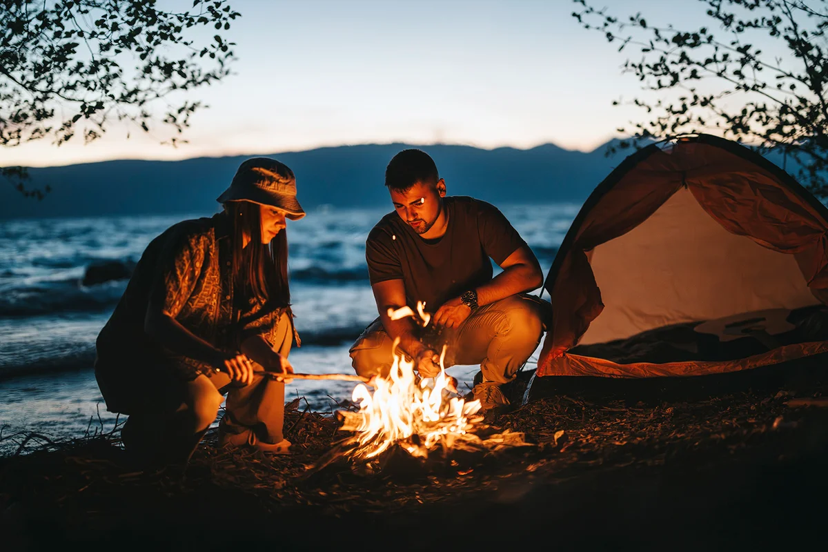 Two friends gather around a bright bonfire near a lake, preparing food while the sunset sets, highlighting the beauty of the great outdoors.