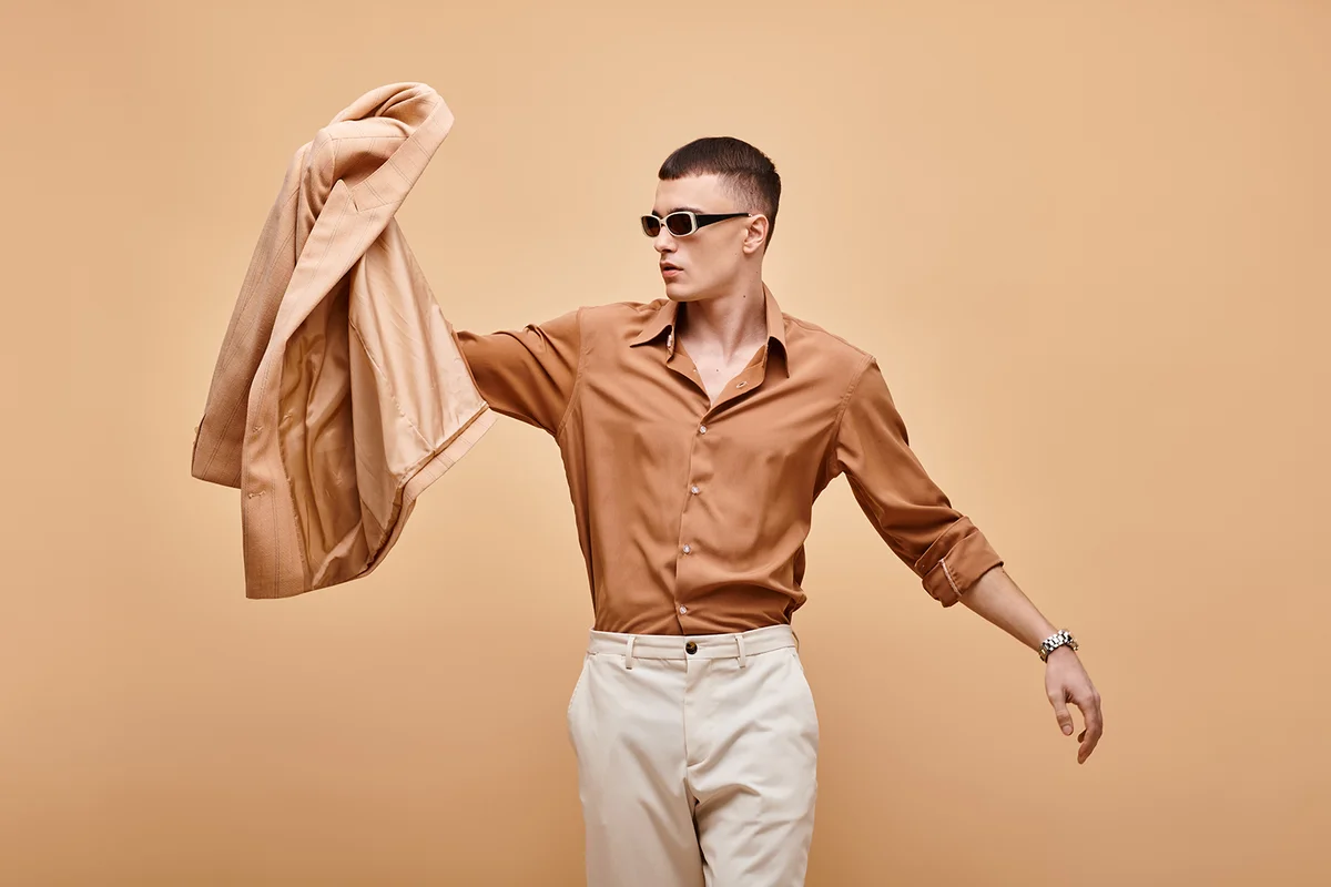 Fashion shot of man in beige shirt with sunglasses and holding jacket in hand on beige backdrop