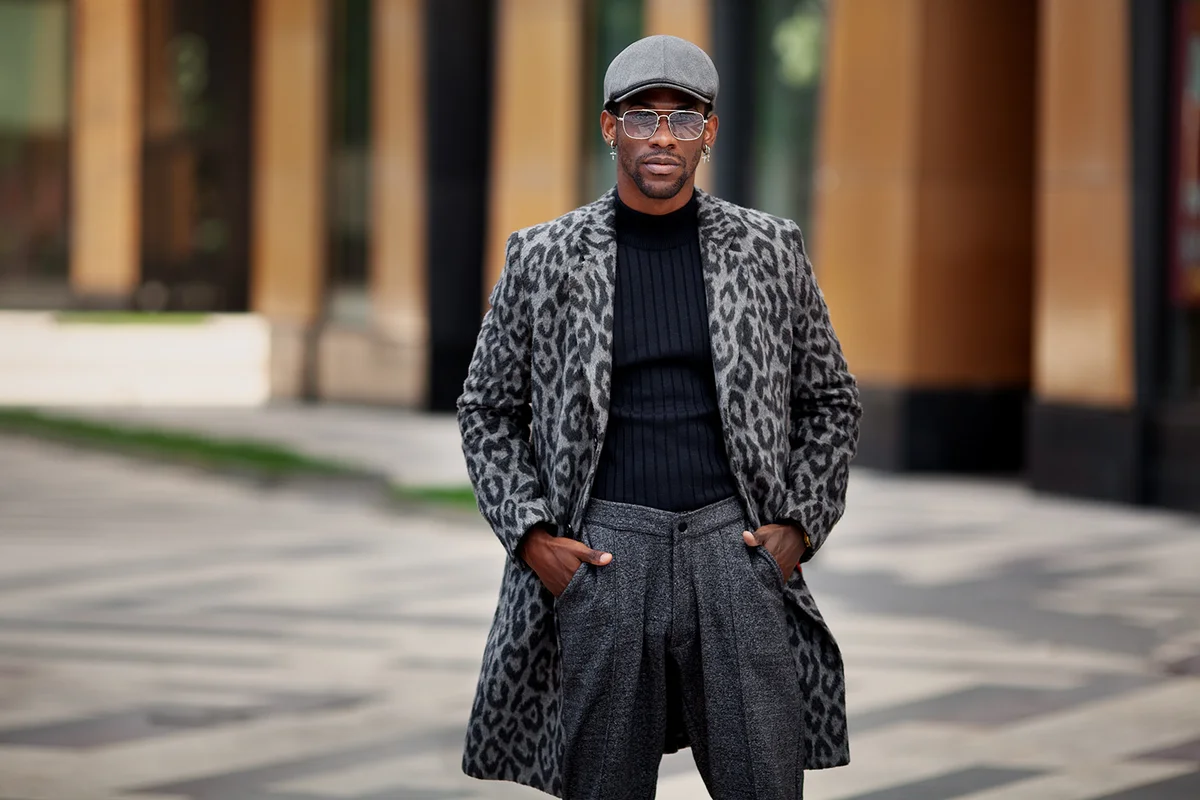 Black young man in fashionable outfit coat with animal print, turtleneck, cap, pants, and glasses, standing on city street and looking at camera