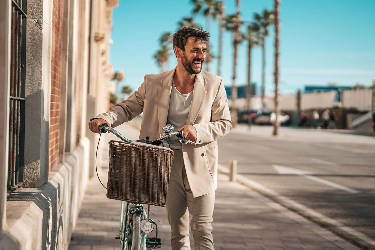 A cheerful Caucasian businessman in smart casual attire rides a bicycle down a sunlit city street