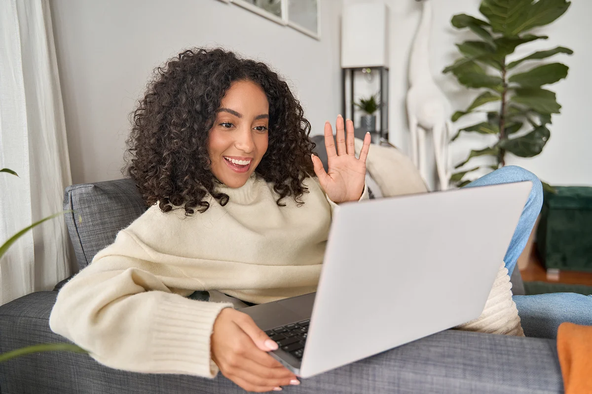 Latin happy smiling curly young woman sitting on couch using laptop at home waving hand