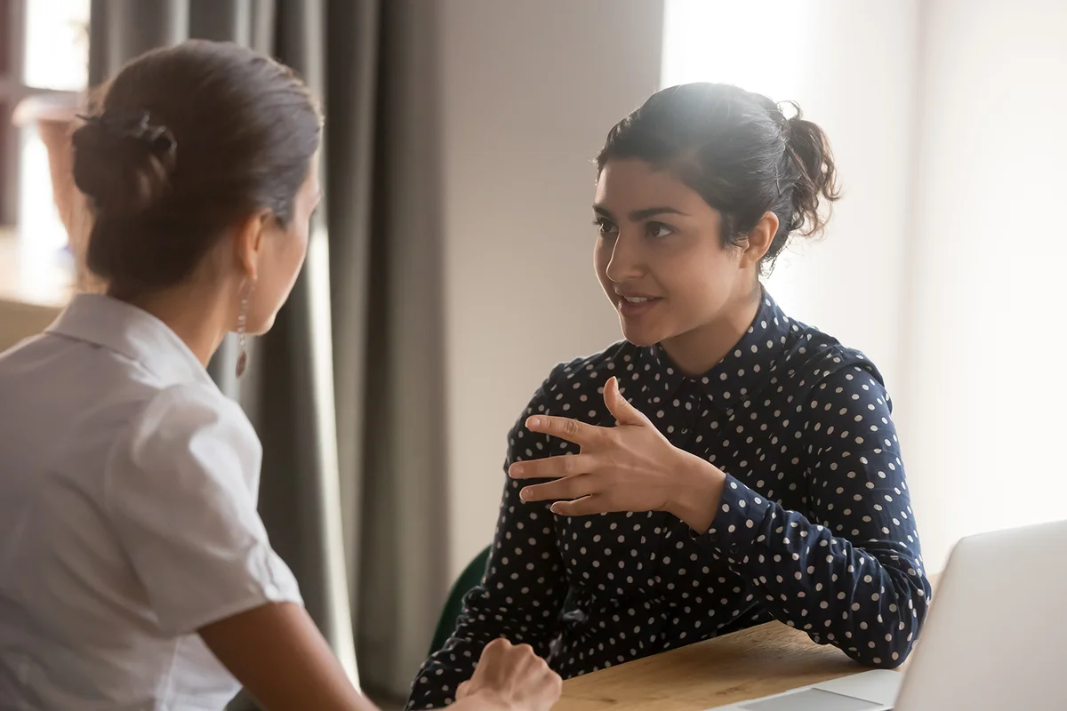 Serious indian mentor teacher worker talk to female colleague teach intern discussing new skills learning sit at work desk