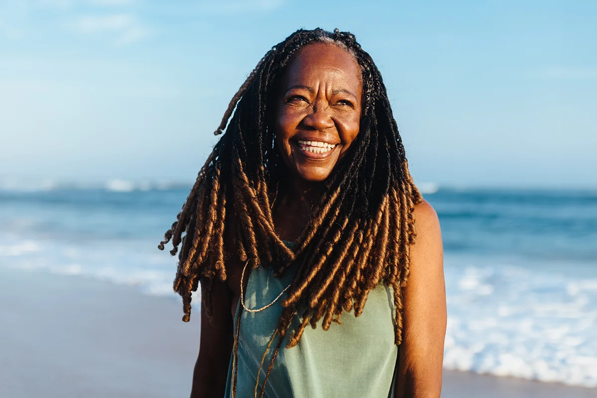Smiling senior woman with braids standing casually on the beach during sunset.