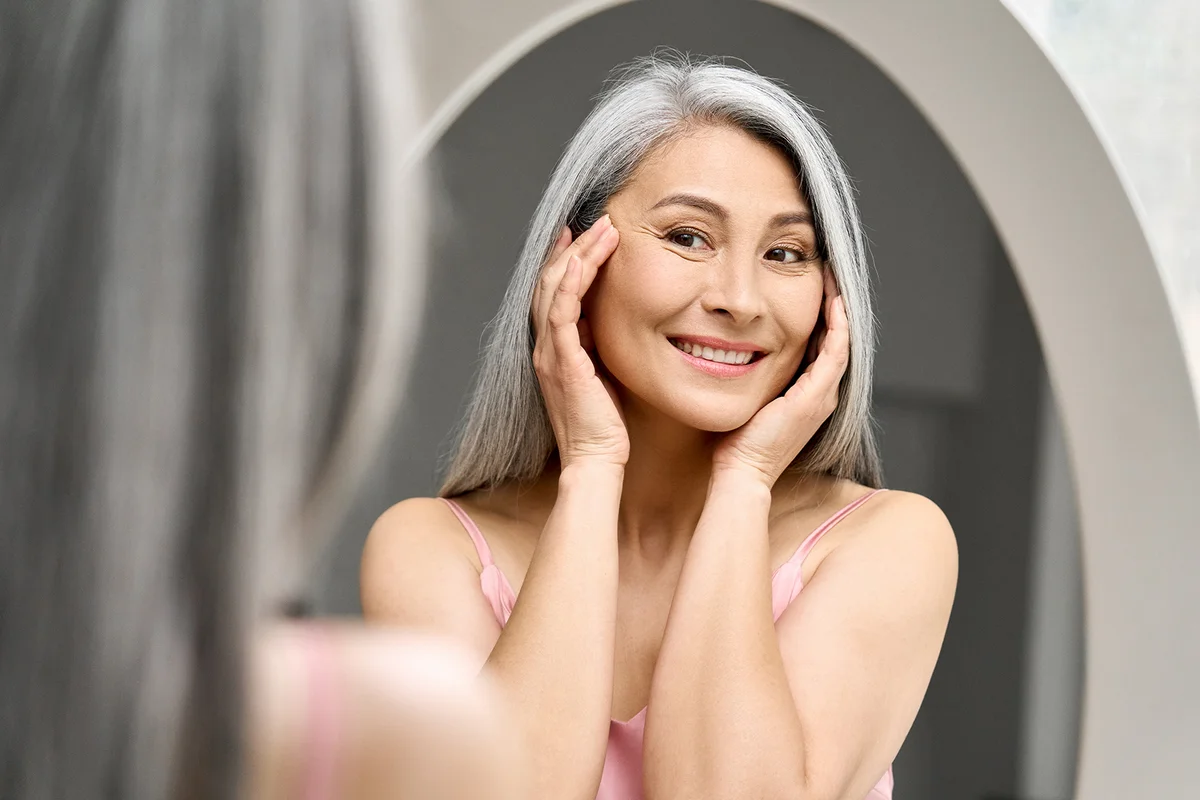 woman with gray hair looking at mirror reflection examining touching face enjoying antiaging beauty treatments