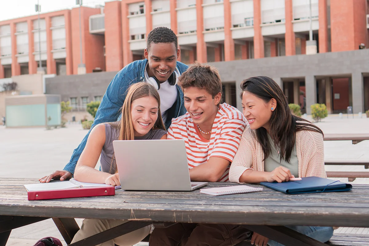 Group multi-ethnic smiling teenage students using laptop while looking information for an academic project outdoors.