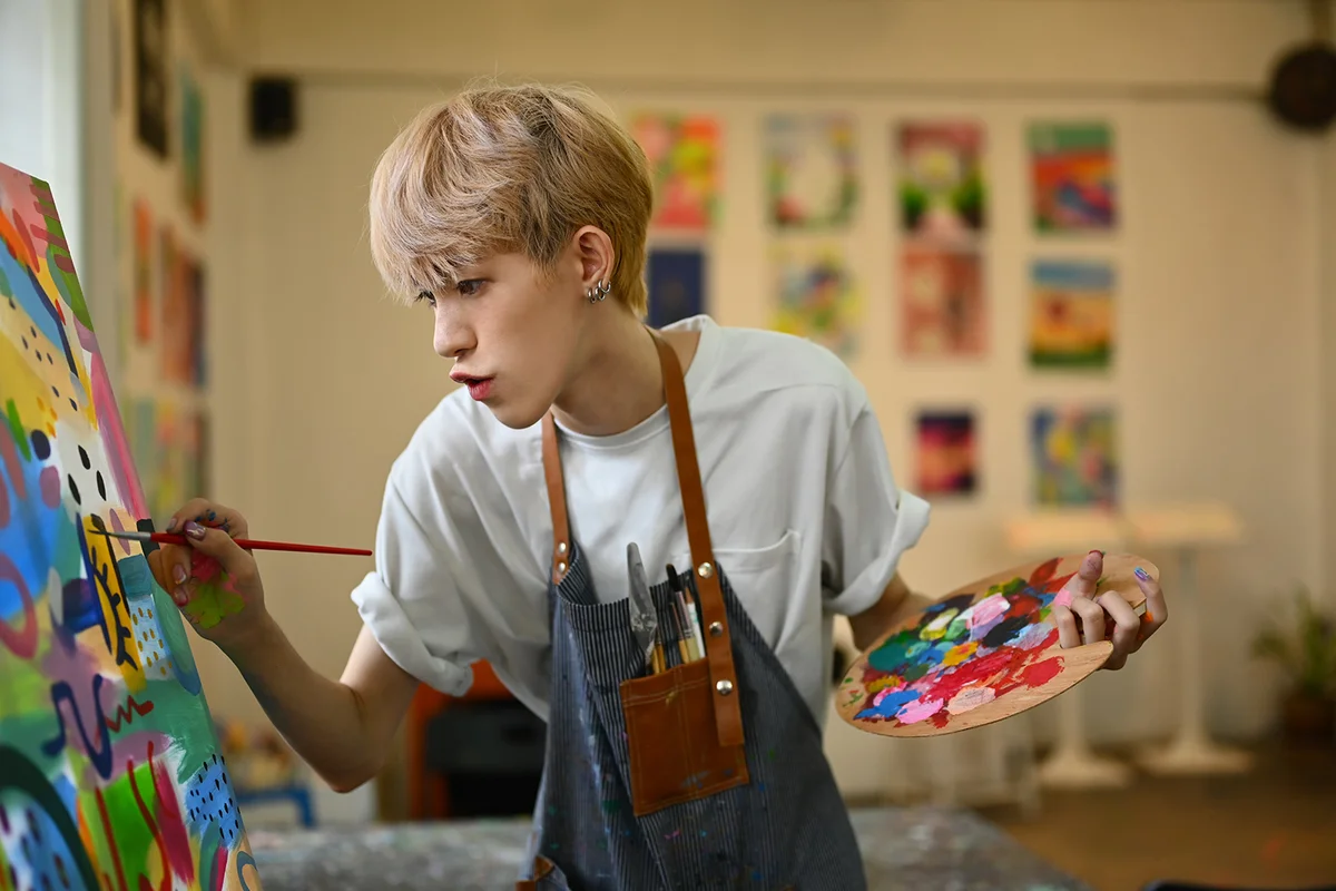 Concentrated teenage asian gay man dressed in apron painting picture with oil paints on canvas in bright art class