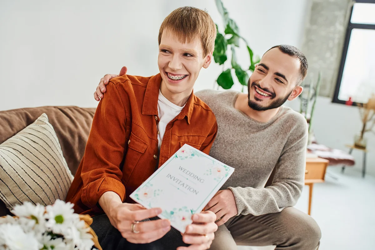 overjoyed couple showing wedding invitation to smiling friend at home