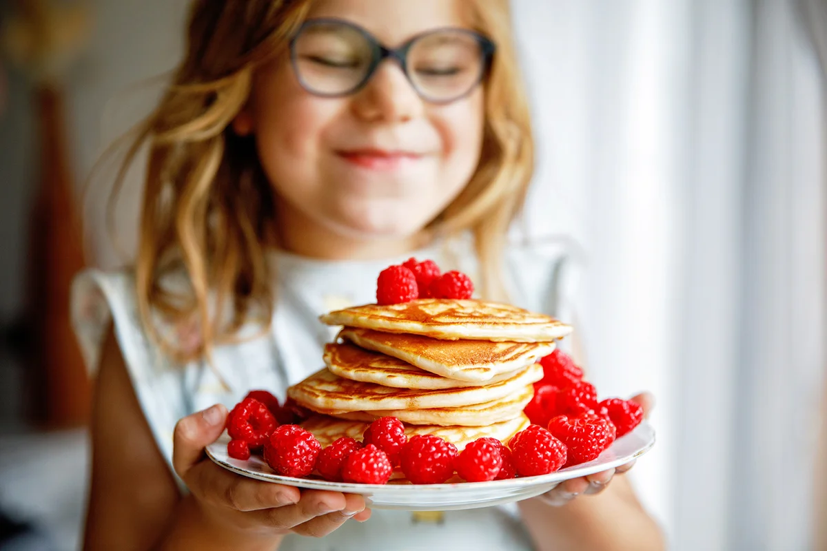 happy preschool girl with a large stack of pancakes and raspberries for breakfast