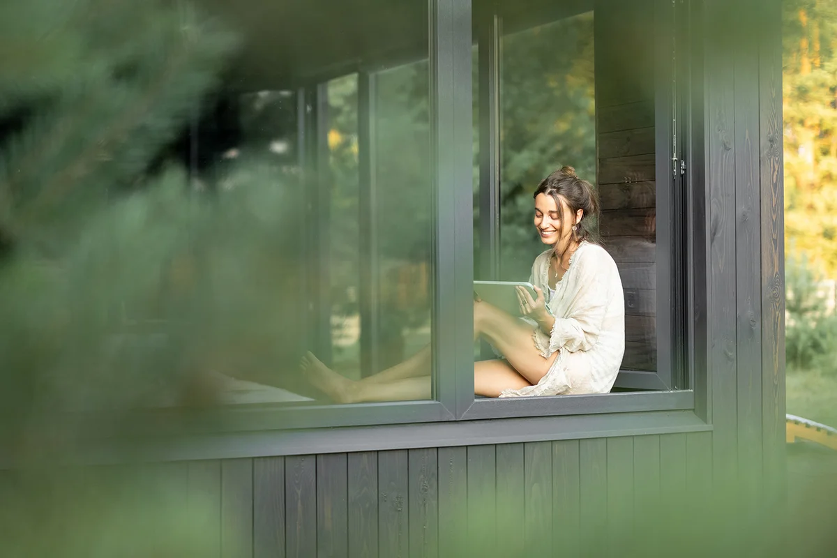 Young woman resting at beautiful country house or hotel, sitting with tablet on the window sill enjoying beautiful view on pine forest.