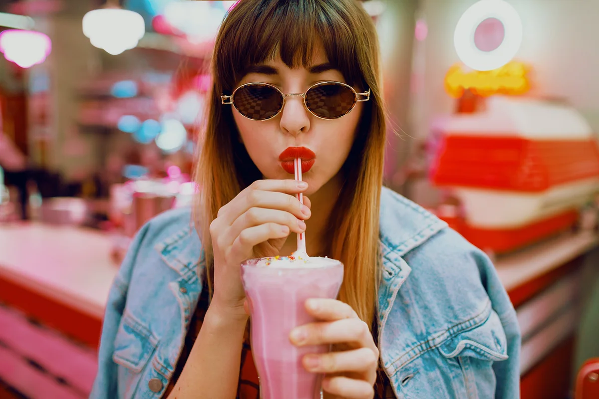 Close up fashion portrait of stylish brunette woman drinking tasty sweet milk shake in retro style American cafe with neon lights.