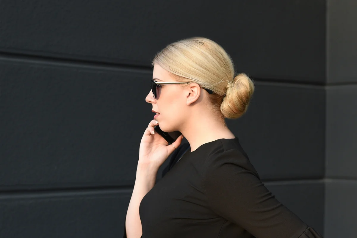 Elegant blonde woman with a bun in a black dress with black sunglasses.