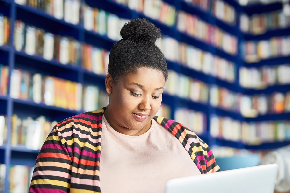 Concentrated portly African-American student girl with hair bun using laptop while doing homework in modern library