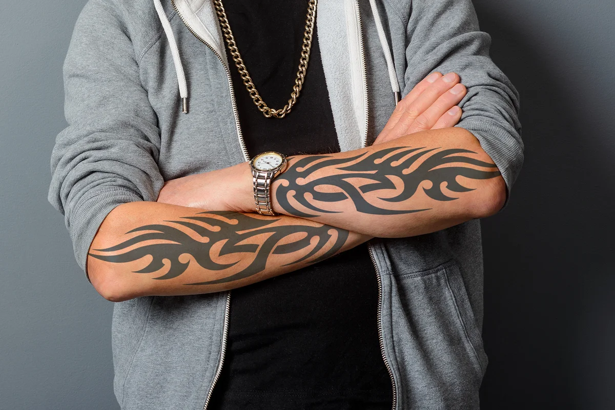 Tattooed arms are crossed, a gold chain hangs over a black t-shirt