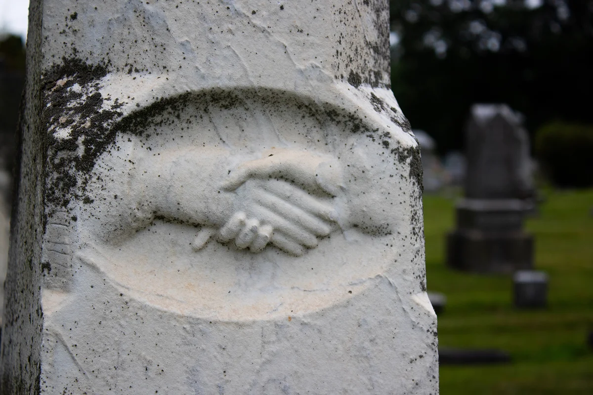 Cemetery headstone closeup of two hands shaking with tombstones in the background