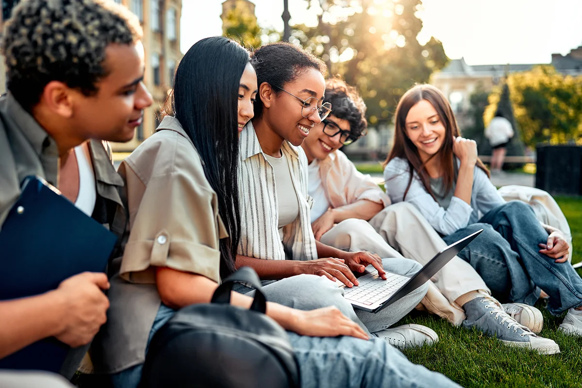 Group of happy diverse students sitting on grass and studying with laptop