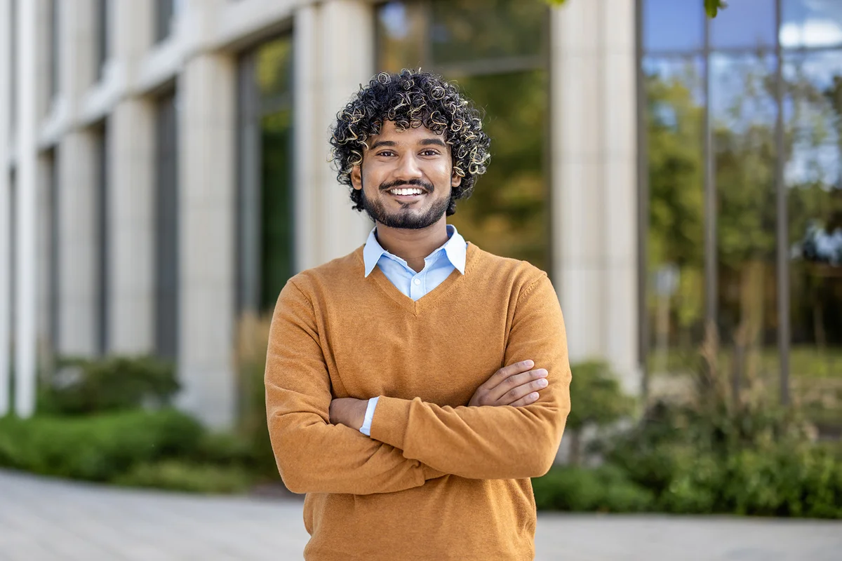Confident young man with curly hair smiling outside modern office building.