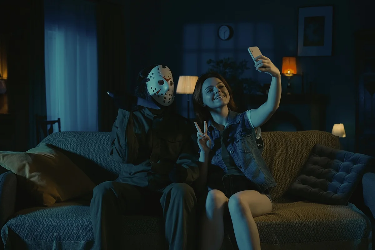 Horror maniac with a knife and young girl sitting on the sofa in the dark apartment room taking selfie on smartphone