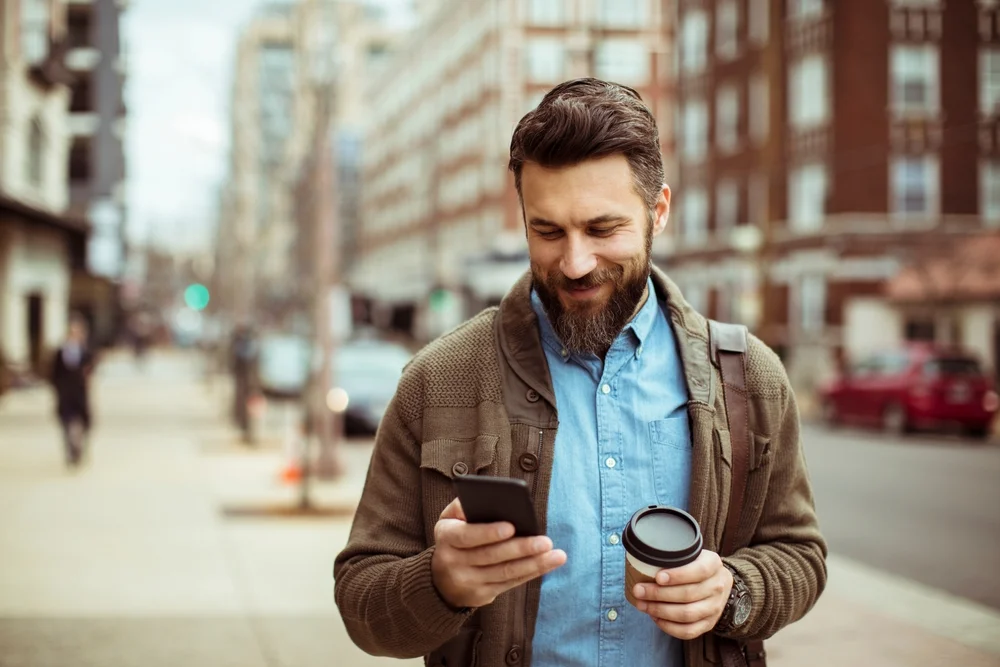 A smiling Man texting on his smartphone and holding a coffee on the city street