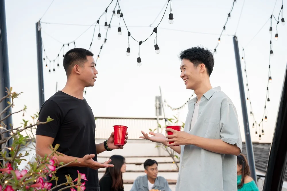 Two happy young Asian male friends enjoy talking while partying on a rooftop bar together