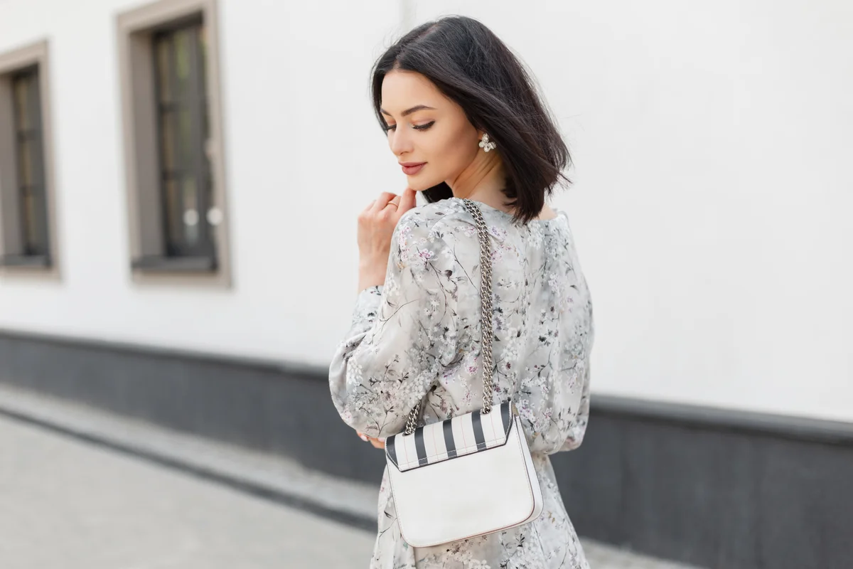 Fashionable young elegant pretty woman model with shorts hairstyle in stylish spring vintage dress with flowers print with fashion white bag walks on the city near a white wall