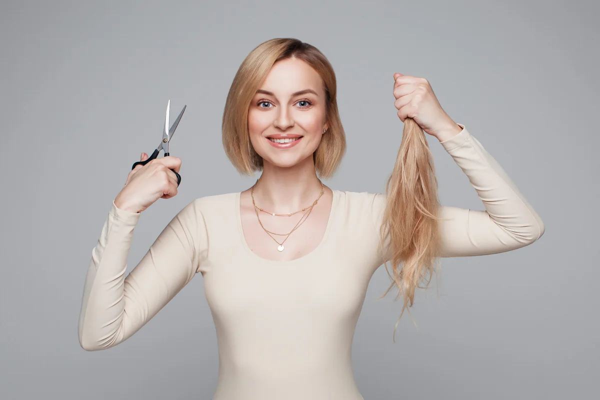 Studio portrait of blonde female cutting her own hair, hairstyle and haircut, lock of hair, scissors in hand, plain background, isolated, changes and hair treatment concept, lifestyle beauty and style