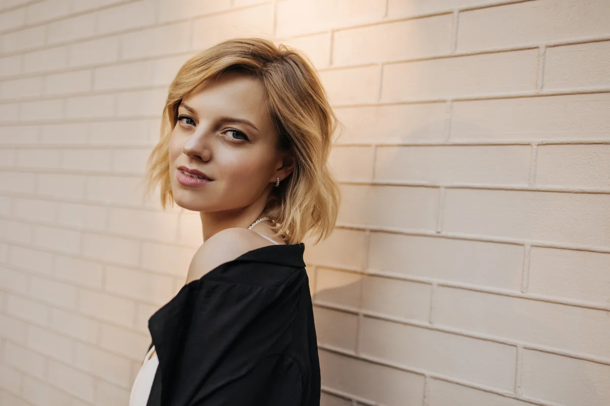 Close-up of young caucasian woman half-sided stands against background of beige wall and looks into camera. Portrait of confident blonde with short shoulder length hair in black shirt.