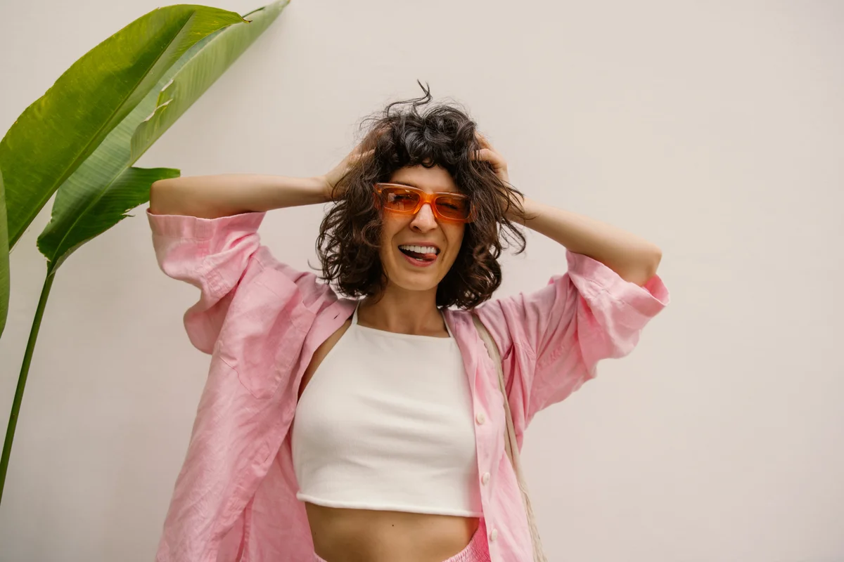 Crazy young caucasian girl grimaces at camera, sticking tongue out, holding hair on white background. Brunette with bob haircut wears glasses, top and shirt in warm weather. Mood, lifestyle, concept.