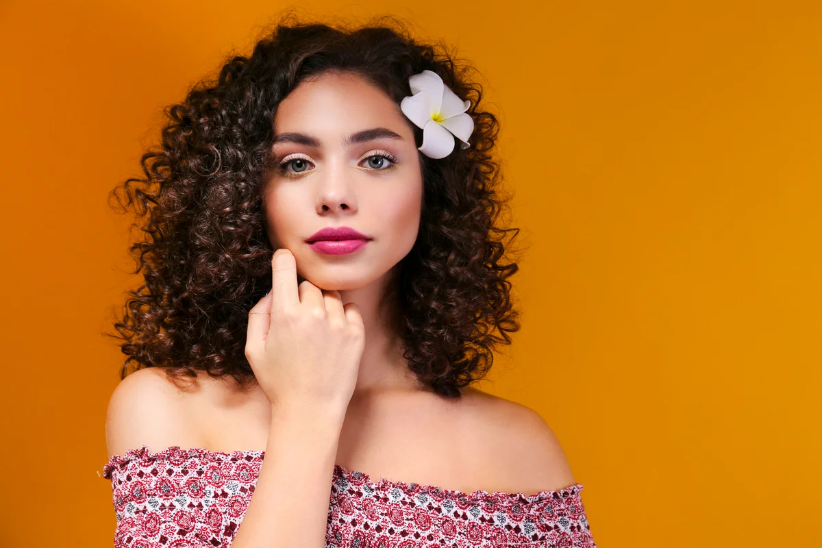 Portrait of happy beautiful woman with long bouncy curles hairstyle and professional make up on, posing over isolated yellow background. Fashion shot of young gorgeous female. Close up, copy space.