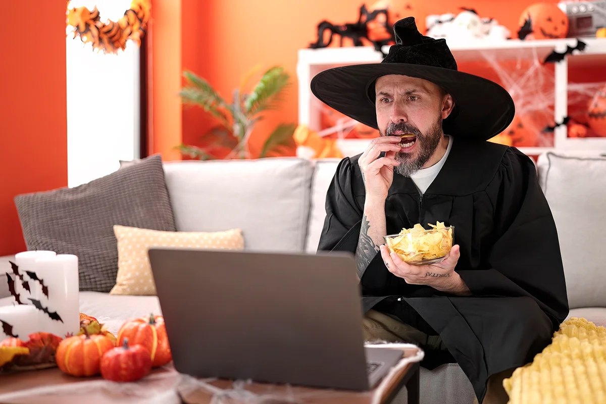 Young bald man wearing wizard costume eating chips potatoes watching movie at home