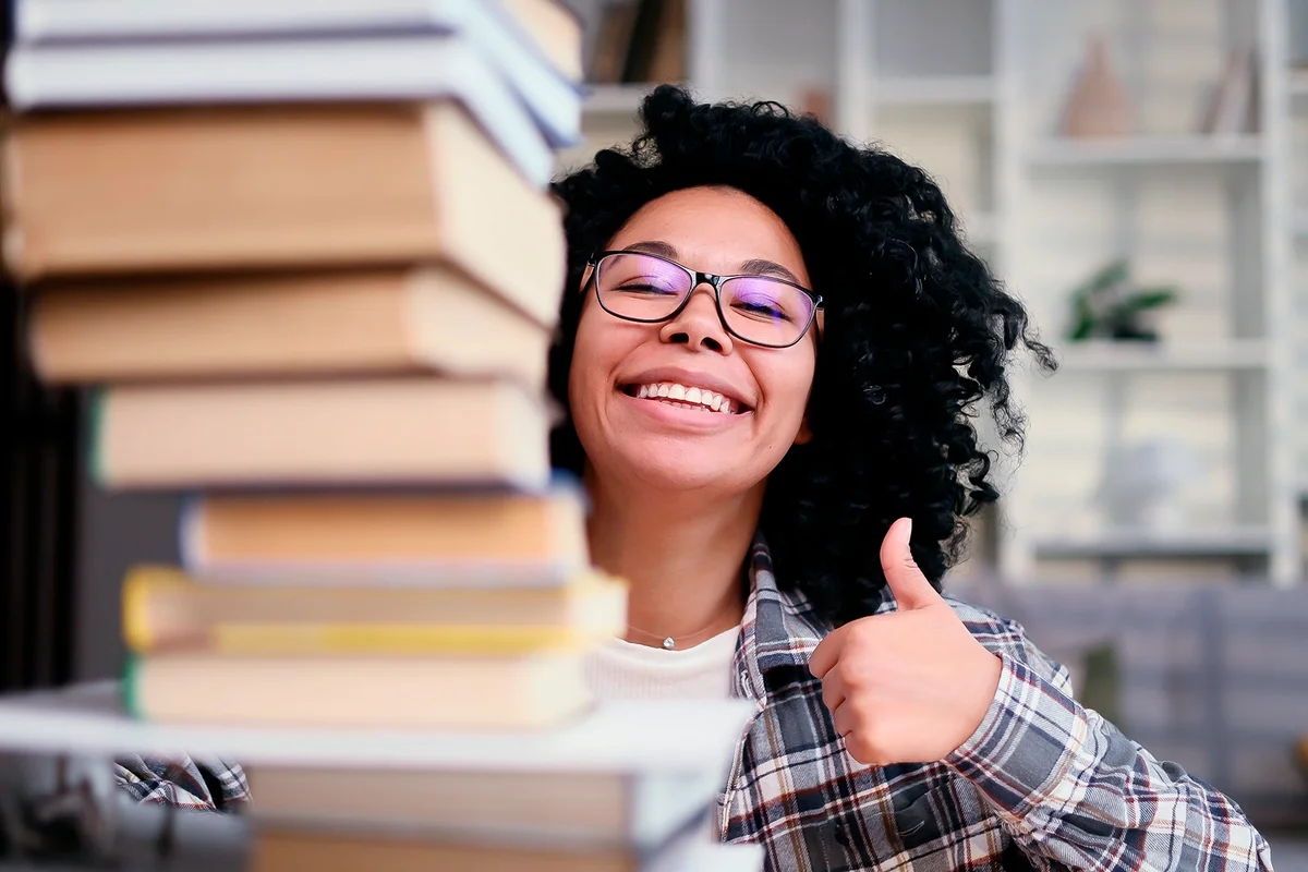 Happy smiling young woman student looking from behind large pile of books