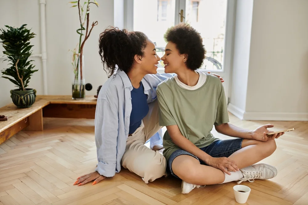 A couple of women look at each other's faces romantically while sitting on the floor at home.