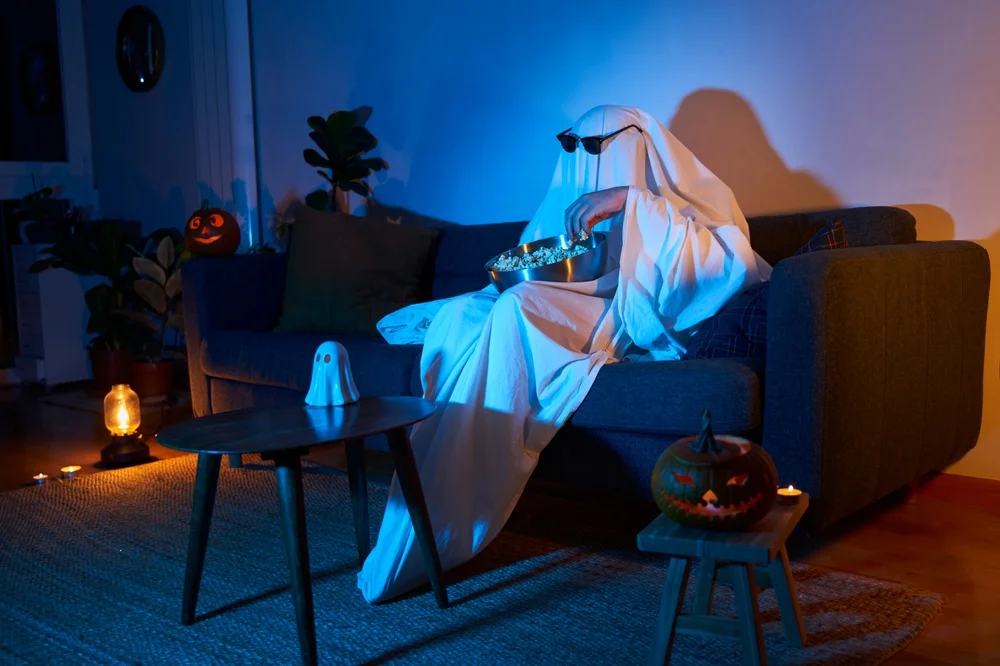 A person in a ghost costume with sunglasses is eating popcorn on the couch at a Halloween-decorated home.