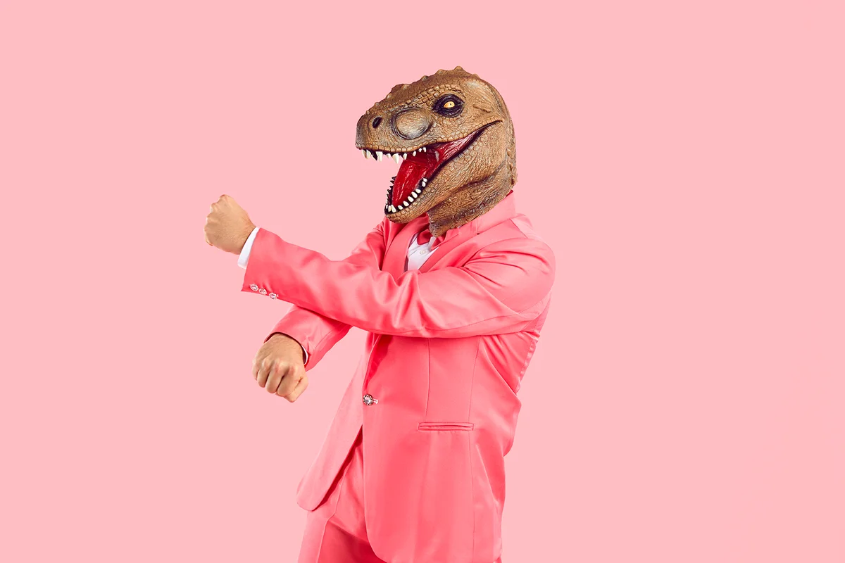 Funny crazy man with mask lizard or dinosaur head dancing isolated on pink background. Man in pink suit puts on animal's head and has fun during costume party. Creative concept for advertising.
