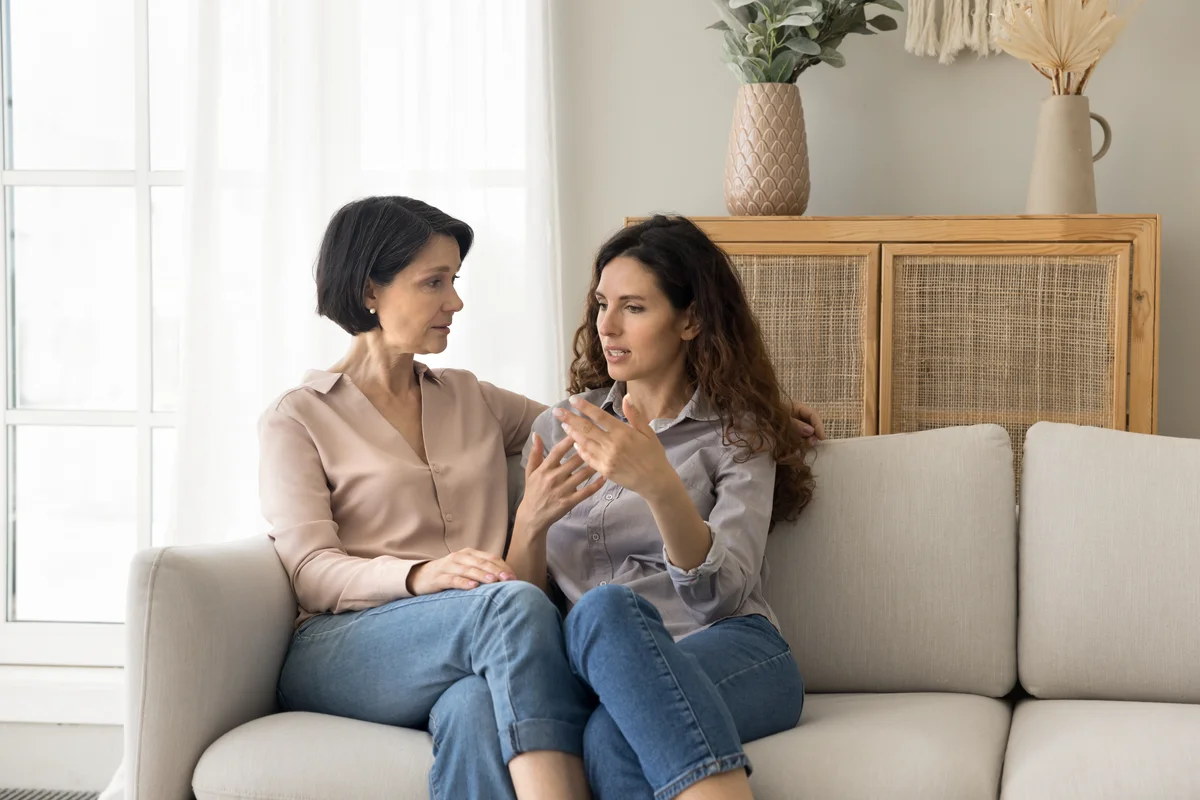 A middle-aged woman sits on a couch, talking with an older woman beside her.