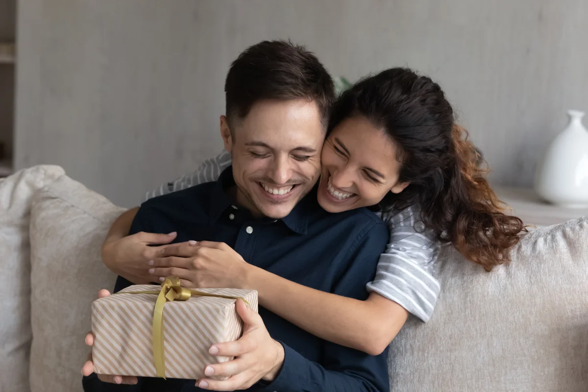 A woman presents her boyfriend sitting on the couch with a gift.