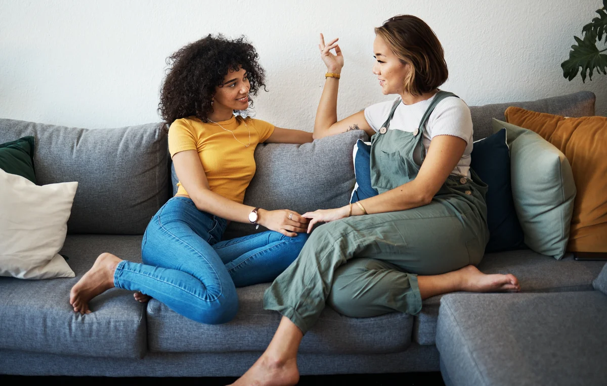Two women sit on a couch holding hands and talking.