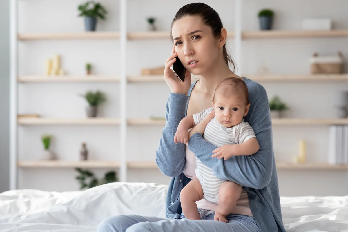 A young mother talks on the phone while holding a baby in her arms.