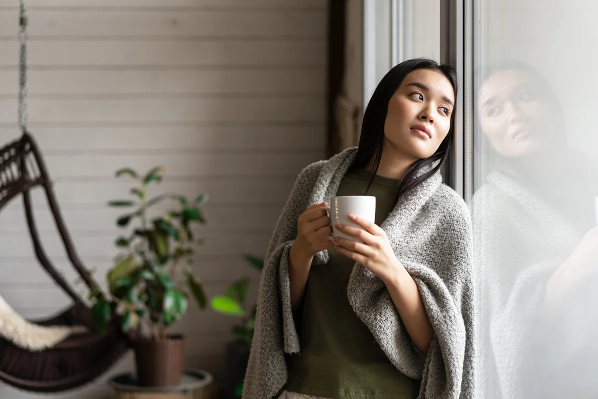 woman wrapped in blanket, leaning on window and looking outside