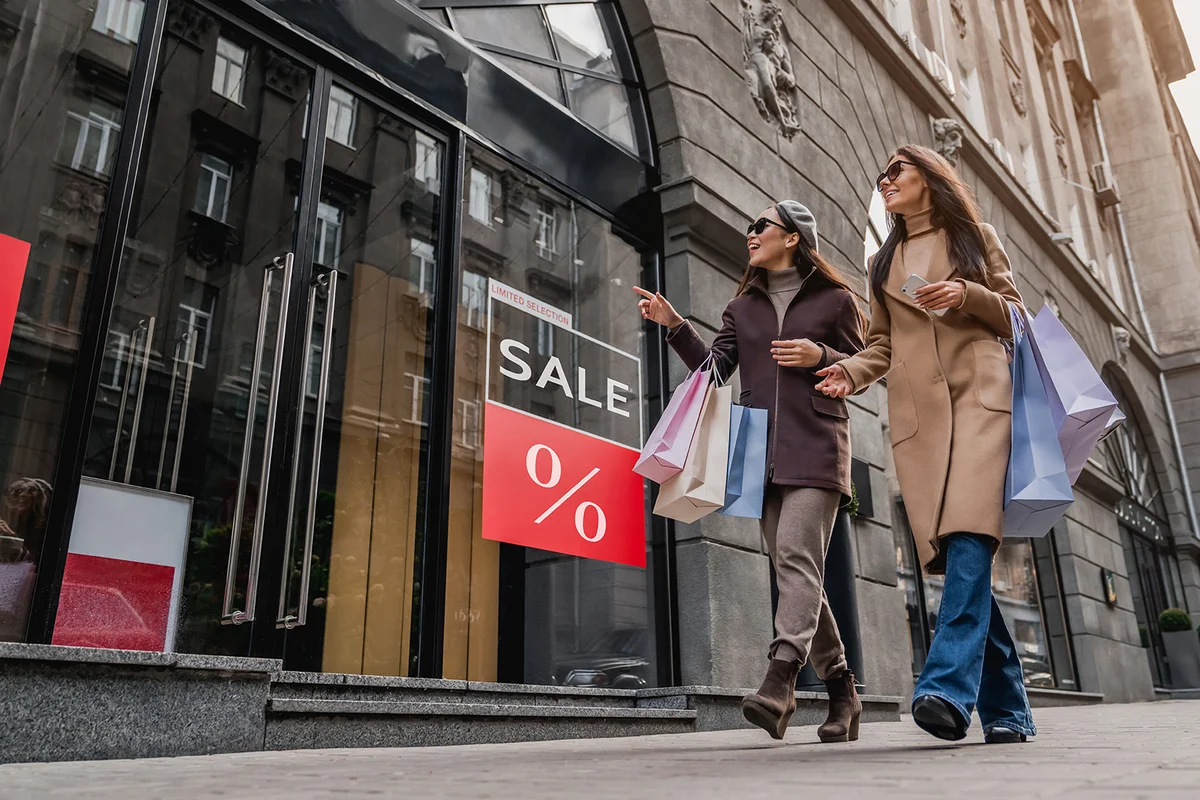 women walking with shopping bag near Sale sign on shop showcase