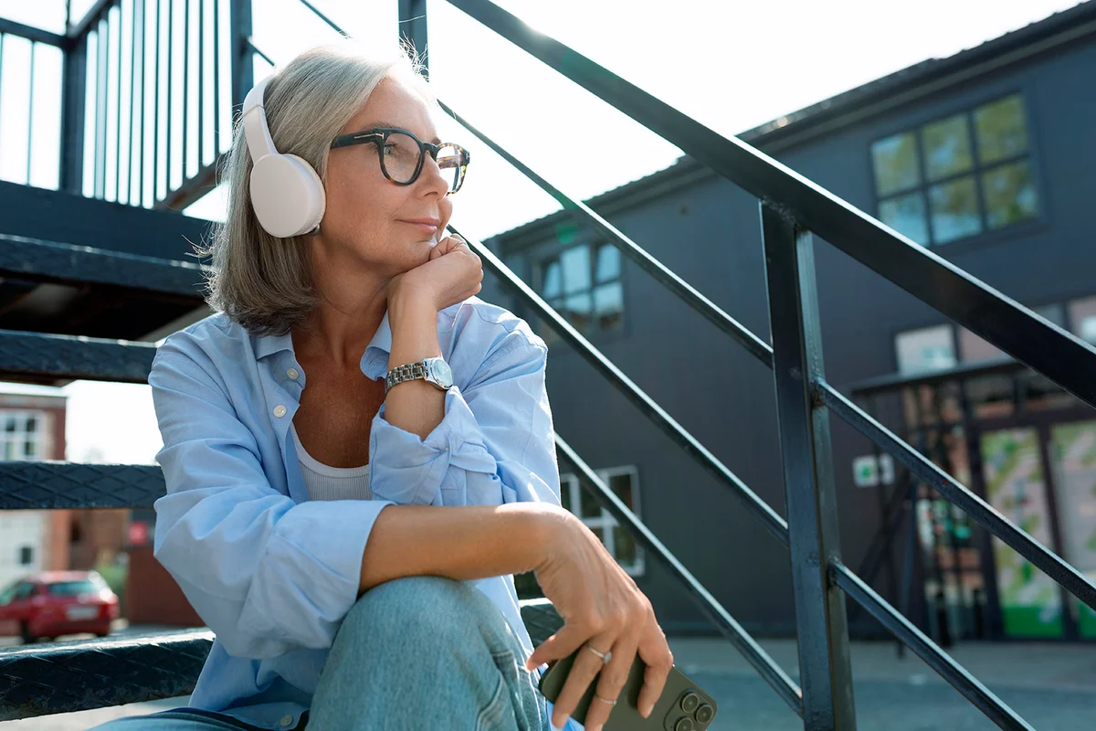 beautiful mature old woman with gray hair dressed relaxes listening to music with headphones in an urban environment